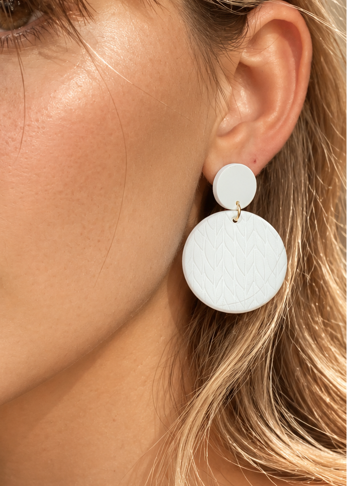 A close-up of a woman wearing a pair of white double-disk drop earrings with a small gold-tone connector, set against a warm, sunlit background.