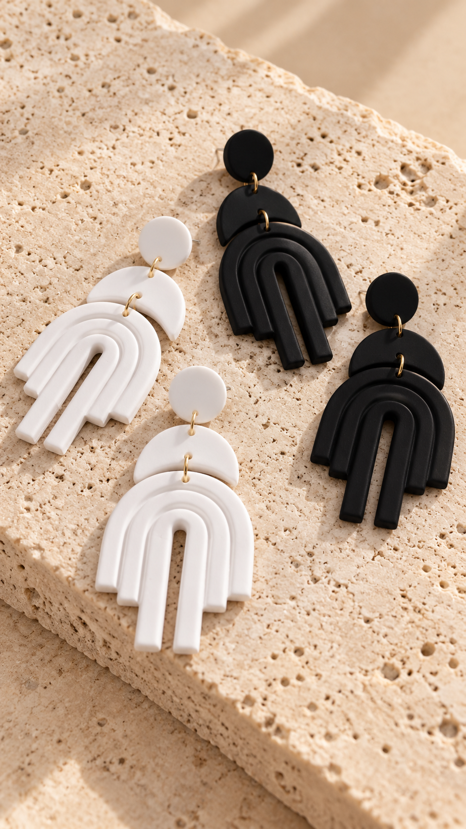 Three pairs of geometric dangle earrings in white and black with gold-tone connectors, displayed on a textured beige stone surface.