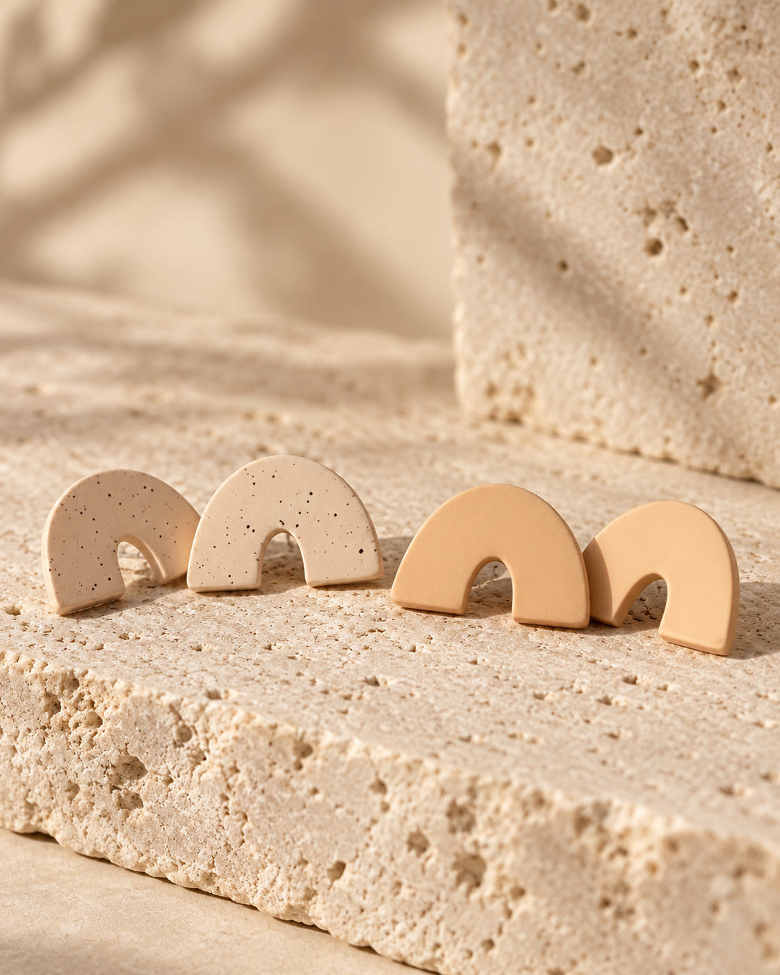 Four small arch-shaped stud earrings in warm neutral tones, including two speckled beige and two solid tan, arranged on a textured stone surface.