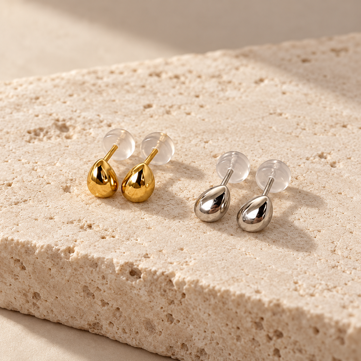 Two pairs of teardrop-shaped stud earrings displayed on a textured beige stone slab. One pair is polished gold with clear silicone backing, and the other is polished silver with clear silicone backing.