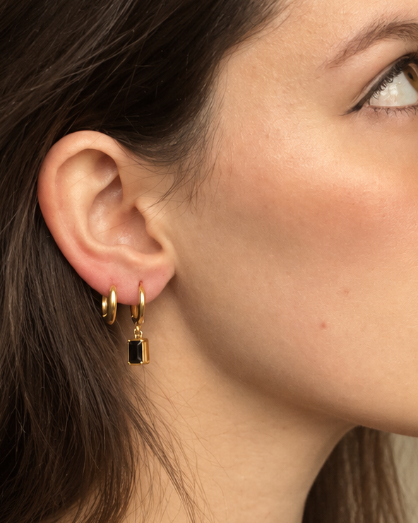A close-up side profile of a woman with dark brown hair wearing a pair of small gold huggie hoop earrings with black rectangular charms, set against a soft beige background.