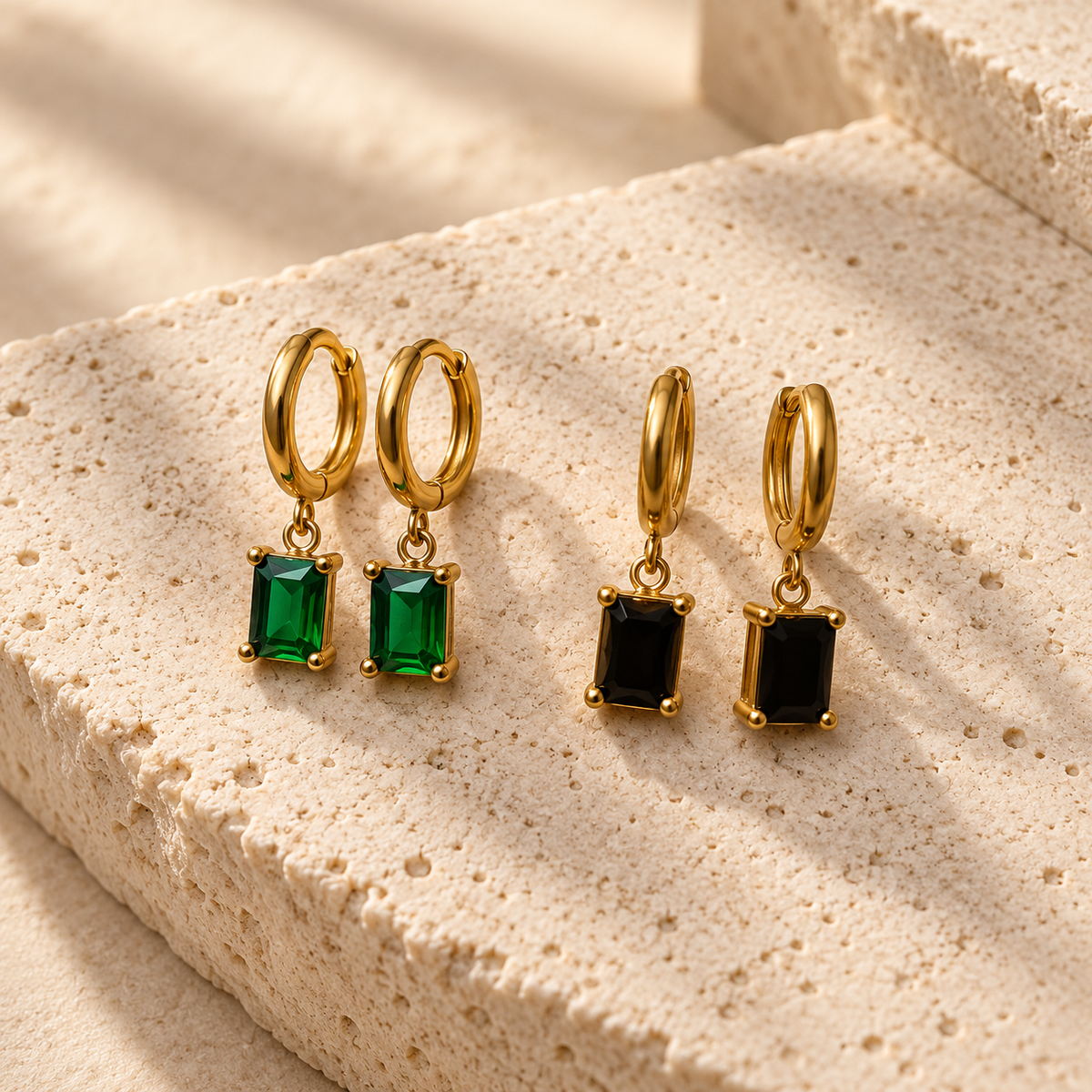 Three pairs of gold hoop drop earrings displayed on a textured beige stone surface. Two pairs feature rectangular green stones, and one pair features rectangular black stones.