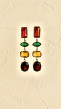 A pair of gold-tone dangle earrings with multiple colored gemstone drops, displayed against a textured beige background.