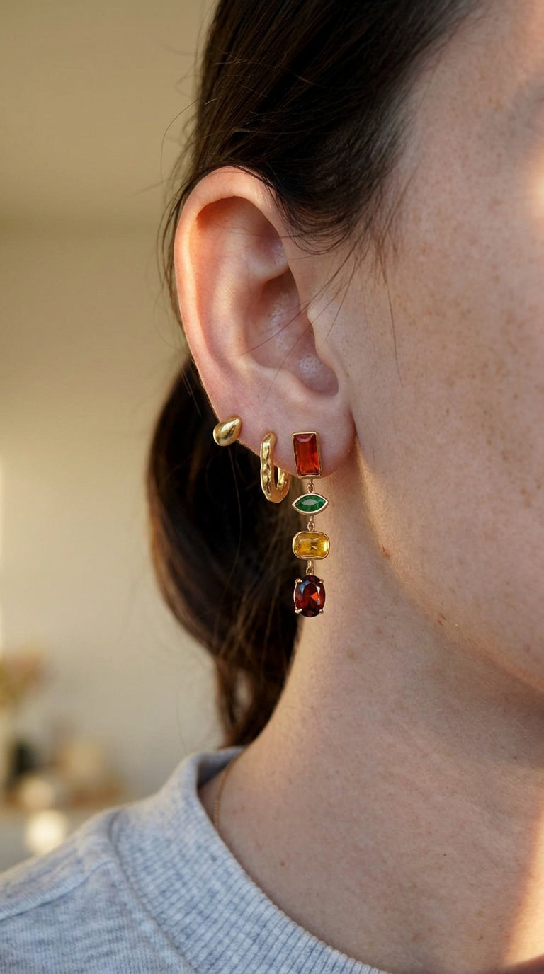 A close-up of a person's ear wearing a stack of gold-toned earrings with colorful gemstone charms, including amber, green, and red stones.