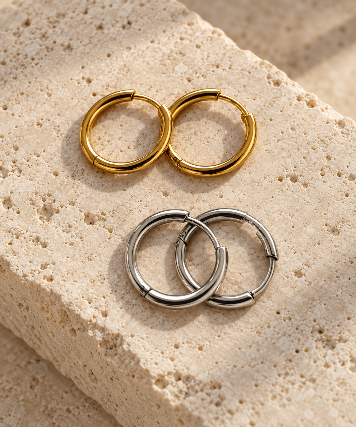 Two pairs of chunky hoop earrings, one in polished gold and one in polished silver, displayed on a textured beige stone surface.