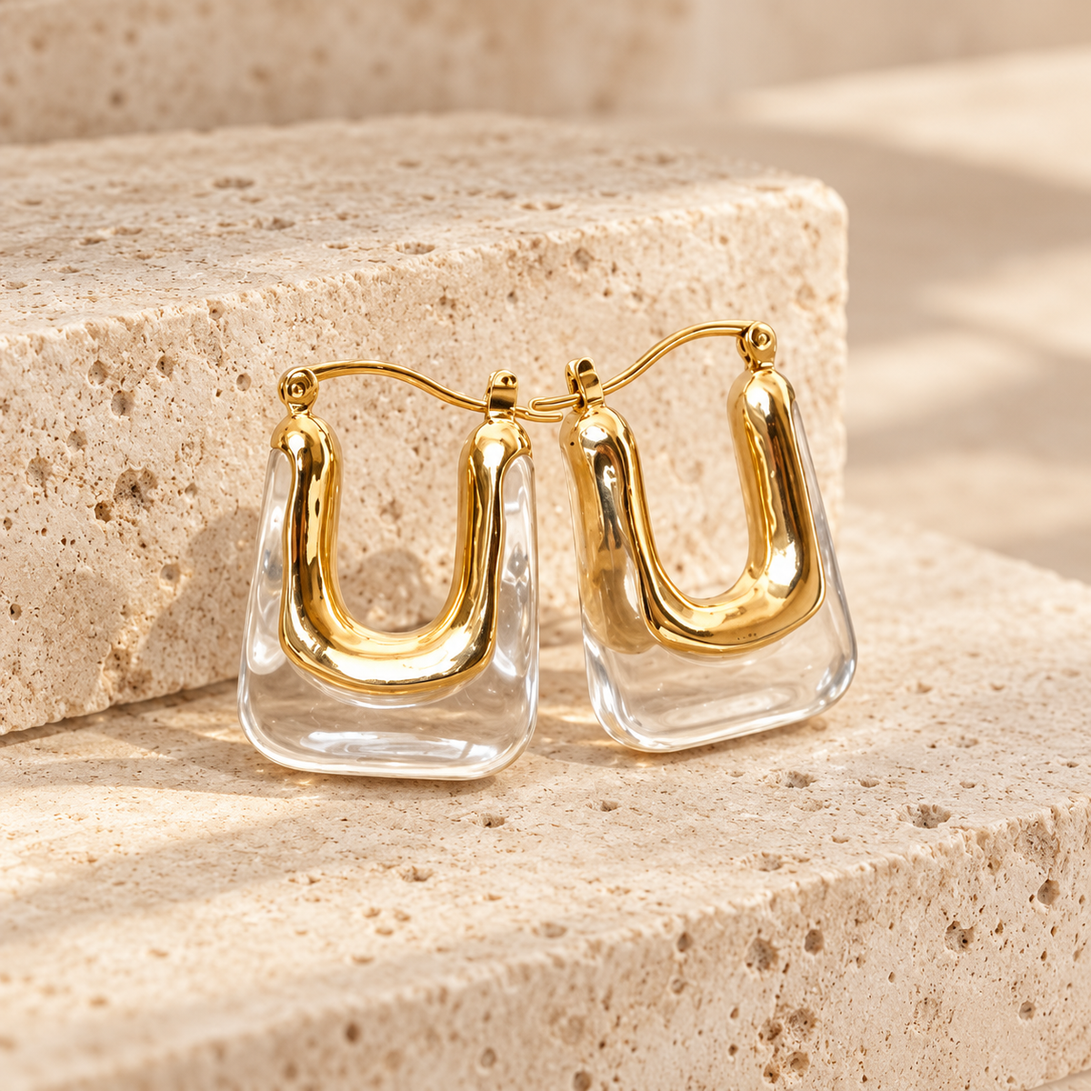 A clear "U" shaped pair of hoop earrings with gold accents are sitting on a light beige stone. 