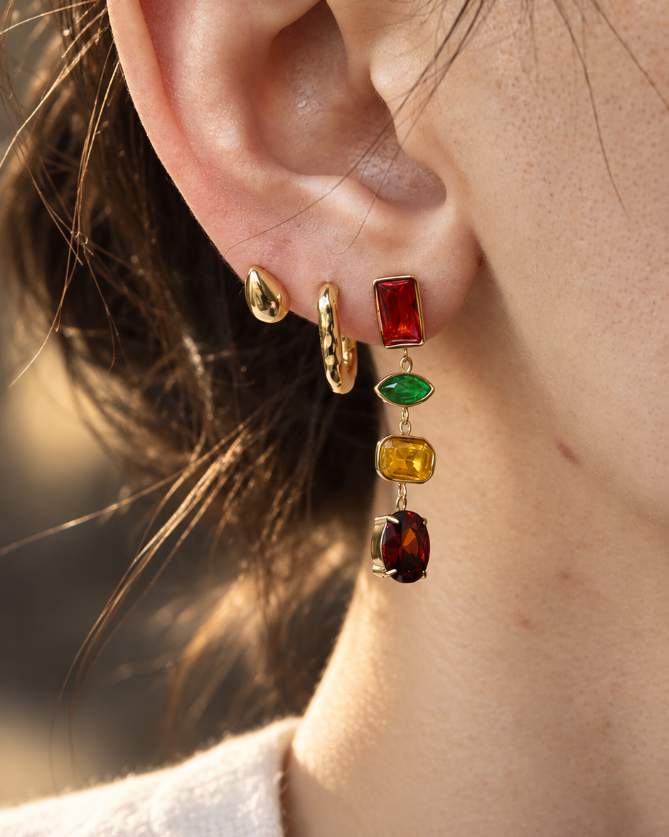 A close-up of a person's ear wearing a stack of gold earrings, including a small gold hoop, a gold teardrop stud, and a dangling multi-colored charm earring with red, green, yellow, and brown stones.