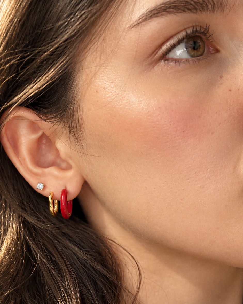 Close-up of a woman's ear wearing a stack of gold hoop earrings, one small silver stud, and a bold red huggie hoop.