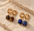 Four gold hoop earrings with dangling square gemstone charms, displayed on textured beige stone blocks. Two pairs feature brown tiger's eye stones, and two pairs feature blue lapis lazuli stones.