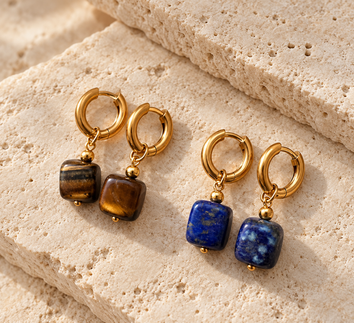 Four gold hoop earrings with dangling square gemstone charms, displayed on textured beige stone blocks. Two pairs feature brown tiger's eye stones, and two pairs feature blue lapis lazuli stones.