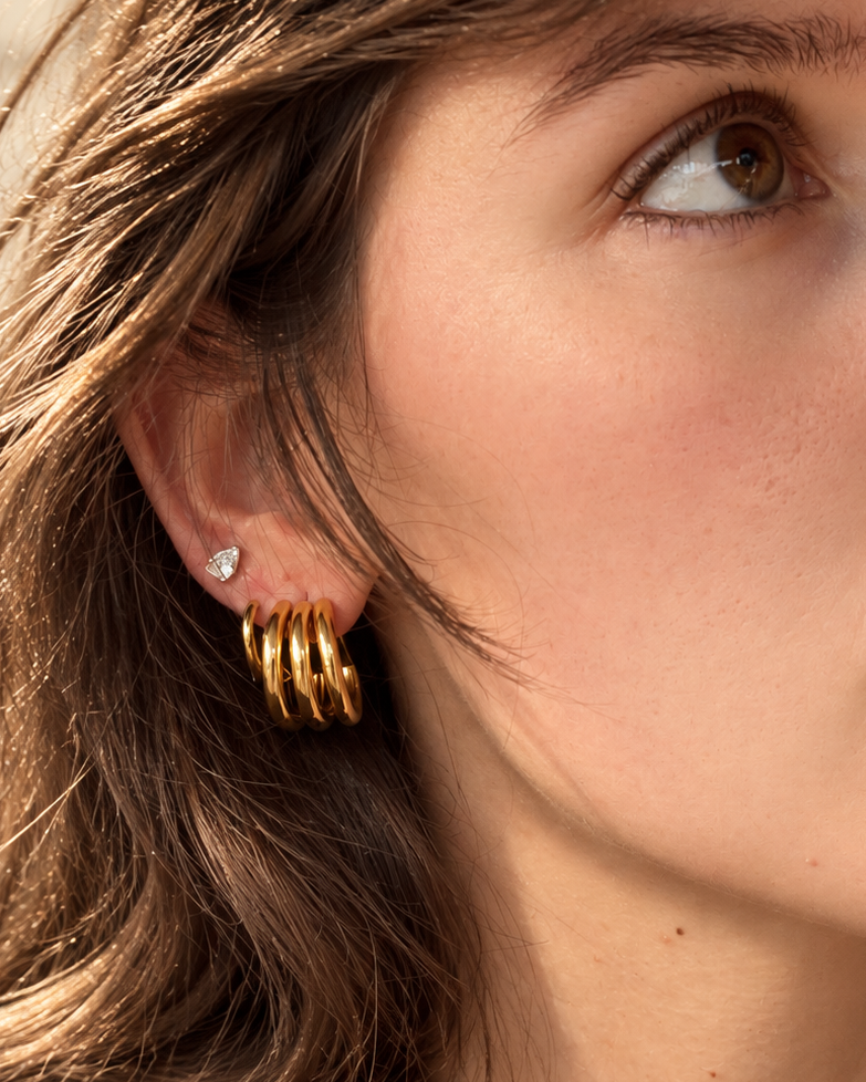A close-up of a woman wearing a pair of gold hoop earrings with a small clear gemstone stud earring, shown in warm natural light.