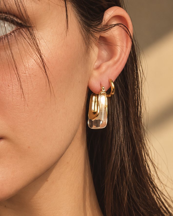 A close-up of a woman wearing a chunky, polished gold geometric hoop earring, with warm sunlight casting soft shadows on her face and hair.