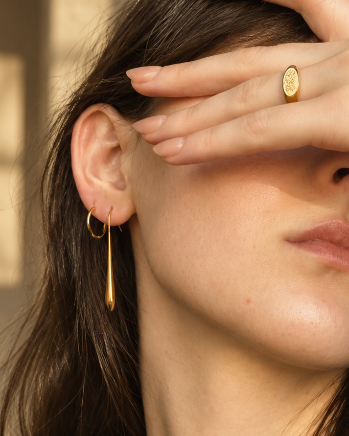 Close-up of a woman wearing gold jewelry, featuring a gold oval signet ring on her finger and a gold huggie hoop earring on her ear, with her hand covering part of her face.