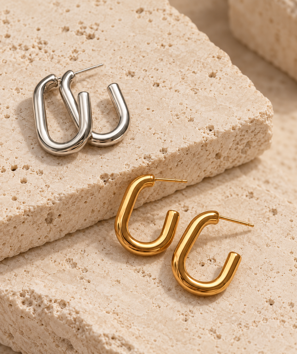 Two pairs of abstract curved hoop earrings, one in polished silver and one in polished gold, displayed on textured beige stone blocks.