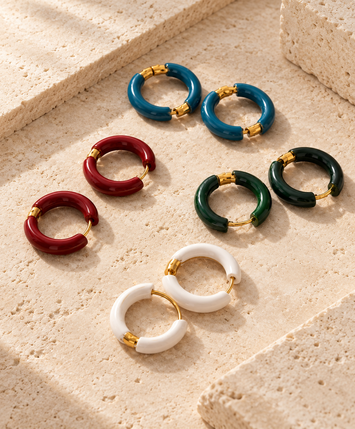 Six pairs of chunky hoop earrings in blue, red, green, and white, each with gold-tone metal accents, arranged on a textured beige stone surface.