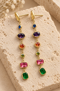 Multicolored seven gemstone drop earrings lay on a light beige stone. 