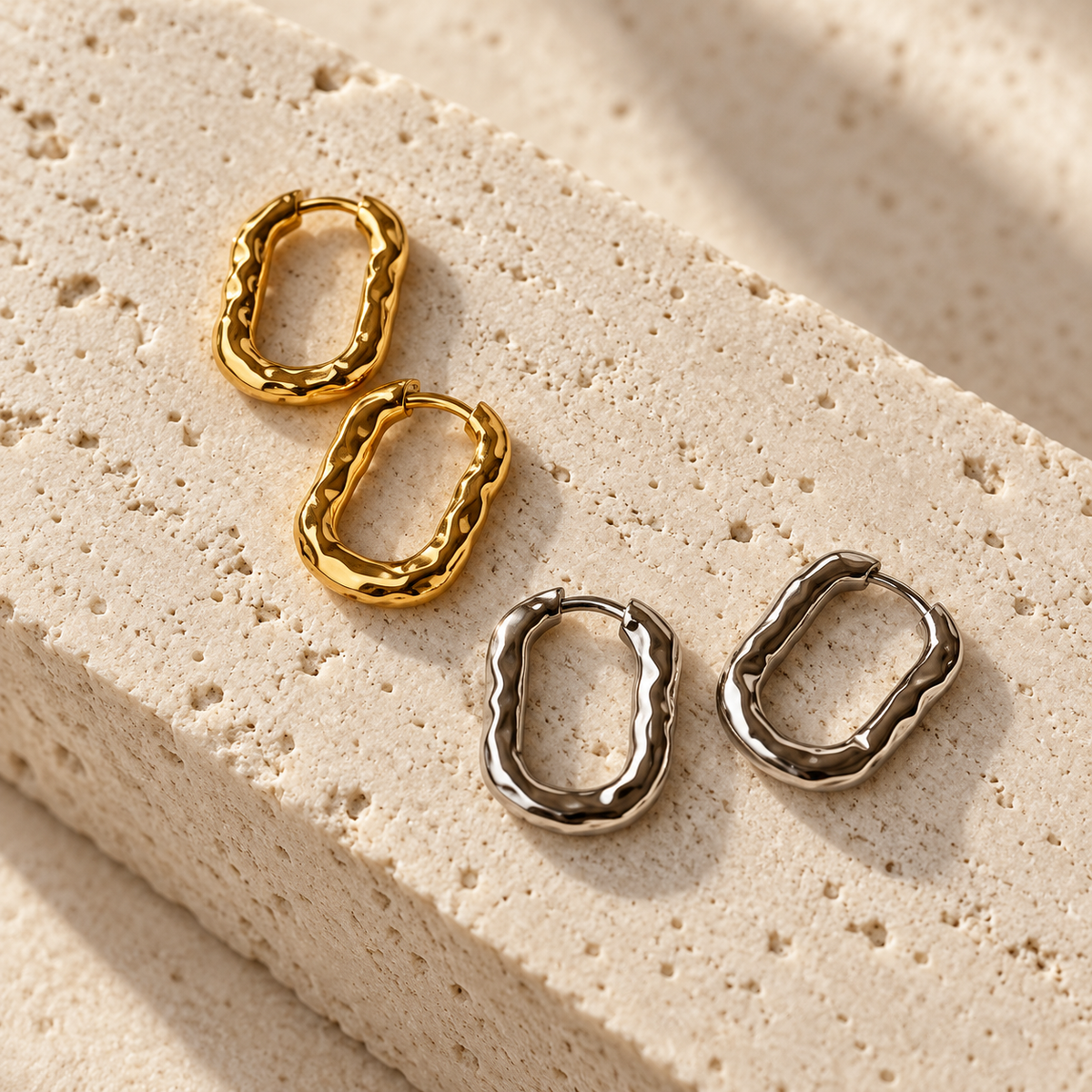 Four textured hoop earrings in gold and silver tones arranged on a beige stone slab with soft shadows.