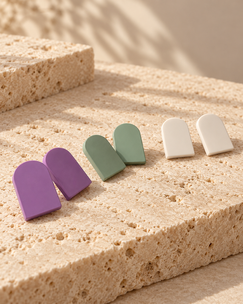 Four dome-shaped stud earrings in purple, sage green, and off-white, arranged on a textured beige stone surface with warm natural shadows.