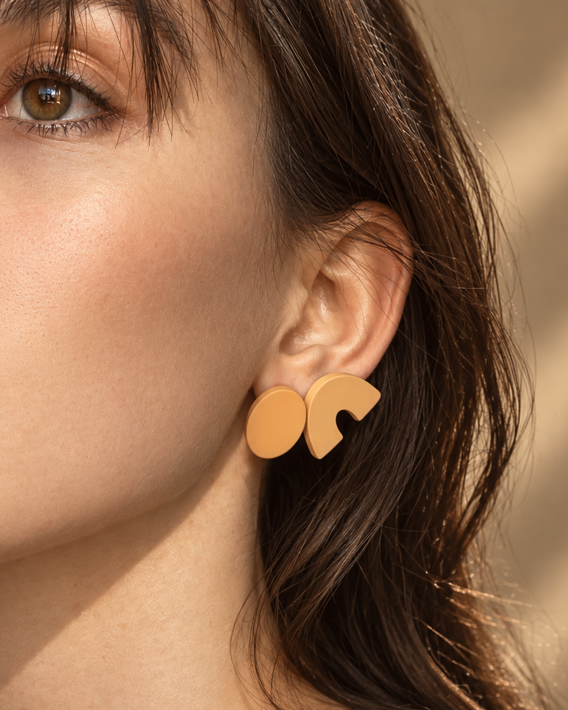 A close-up of a woman wearing a pair of gold geometric stud earrings, with warm sunlight casting soft shadows on her face and neck.