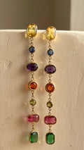 A pair of gold dangle earrings with multiple colorful gemstone beads, hanging from a textured off-white display stand.