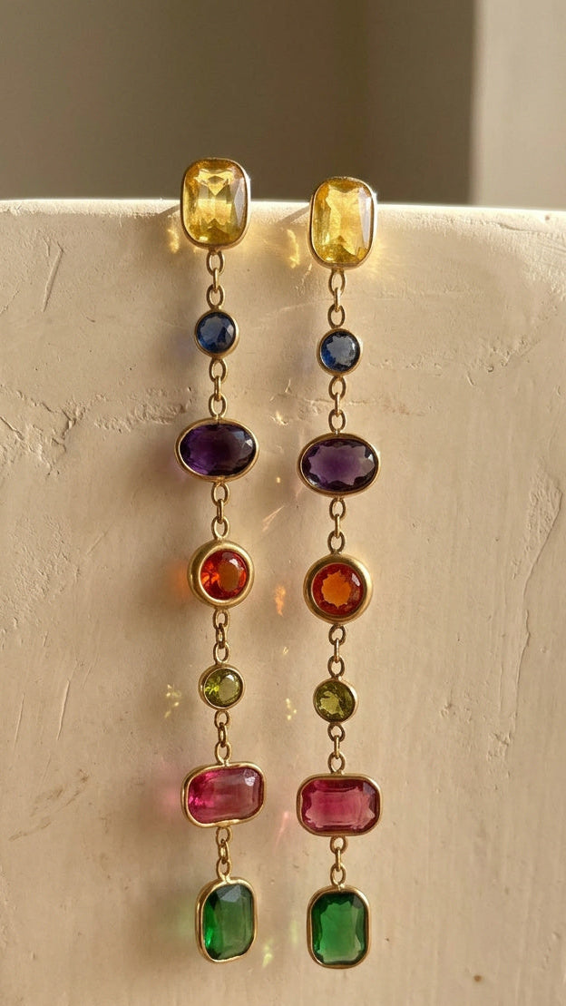 A pair of gold dangle earrings with multiple colorful gemstone beads, hanging from a textured off-white display stand.