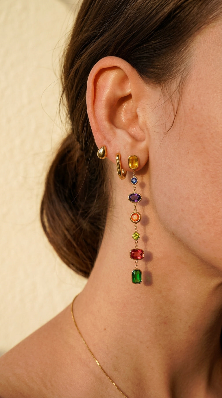 A close-up of a woman's ear wearing a long, multi-colored drop earring with gold-toned settings and colorful gemstone beads, alongside a small gold hoop and a gold teardrop stud.