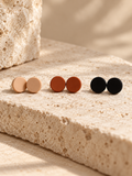 Three pairs of round stud earrings in neutral and earthy tones, displayed on a textured beige stone surface with a warm, sunlit background.
