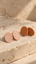 Two oval stud earrings displayed on a textured beige stone surface in warm natural light. Two earrings are light peach with small dark speckles, and one pair is solid terracotta orange.