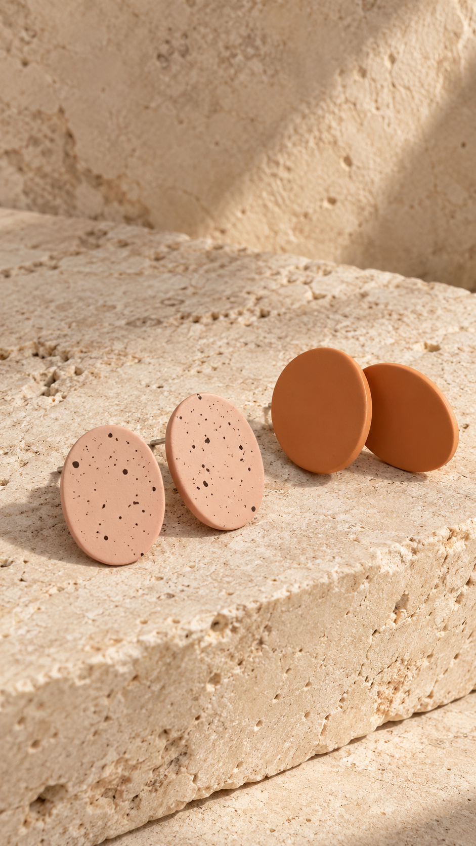 Two oval stud earrings displayed on a textured beige stone surface in warm natural light. Two earrings are light peach with small dark speckles, and one pair is solid terracotta orange.