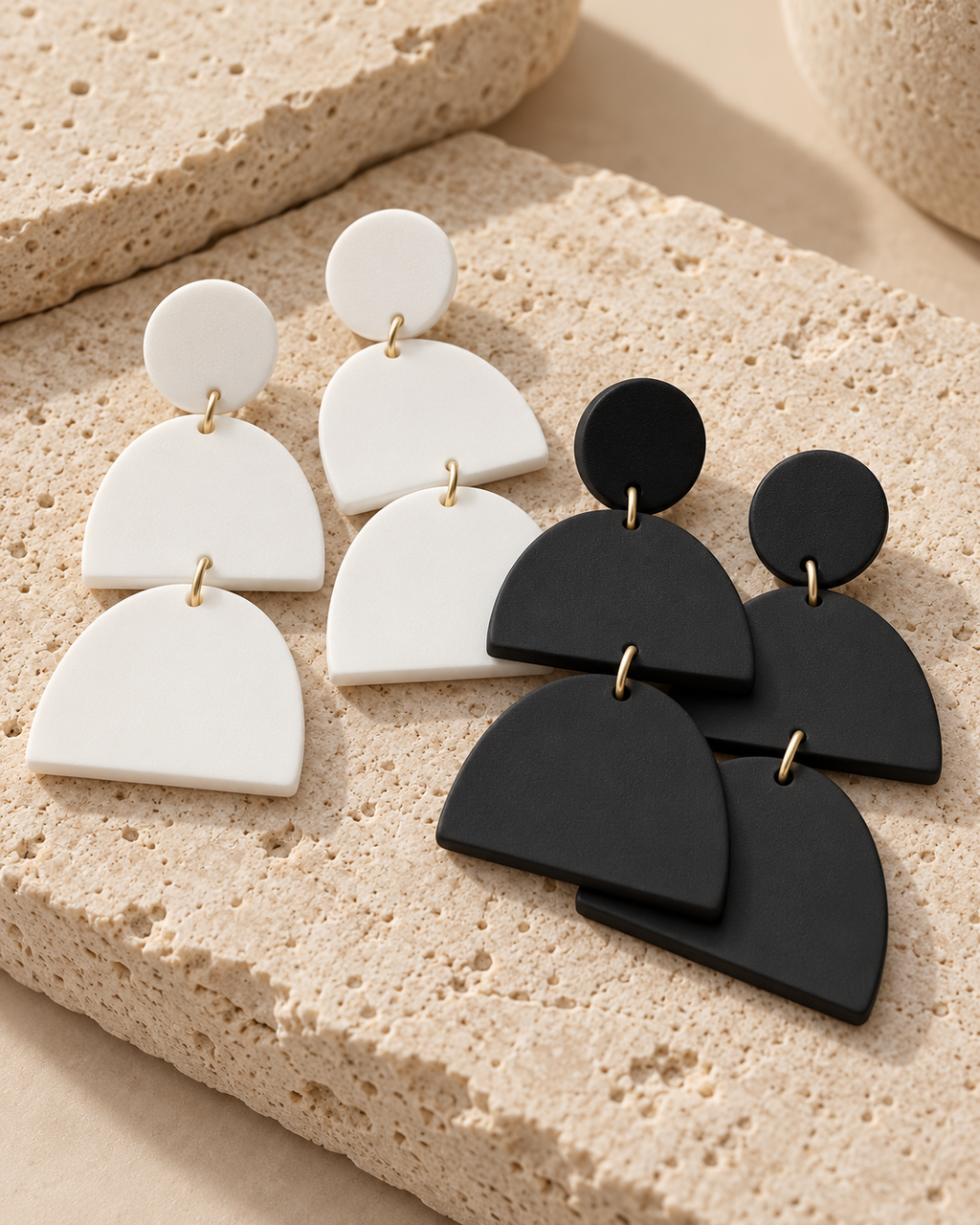 Three pairs of geometric dangle earrings with gold-tone connectors, featuring a small round disc, a semi-circular petal shape, and a larger arch-shaped piece. Two pairs are matte white and one pair is matte black, displayed on textured beige stone blocks.