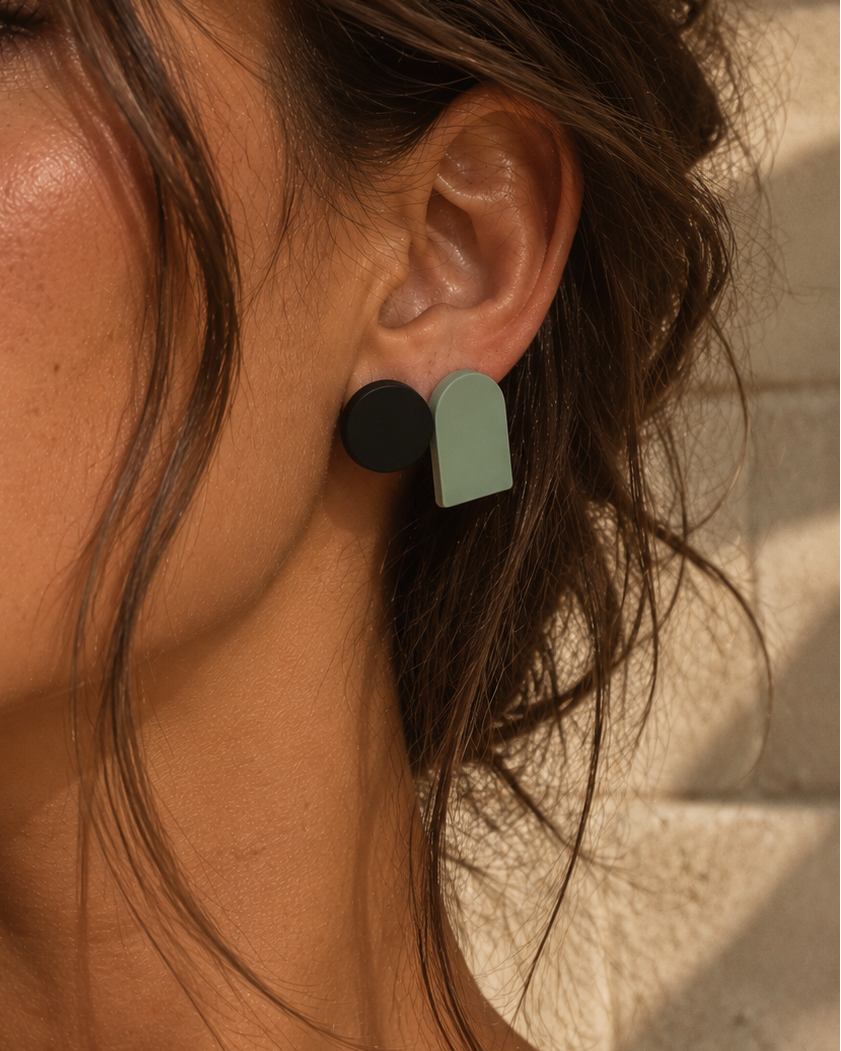 A close-up of a woman's ear wearing a two-tone geometric stud earring with a matte black circle and a light sage green arch shape, set against a warm beige stone wall.
