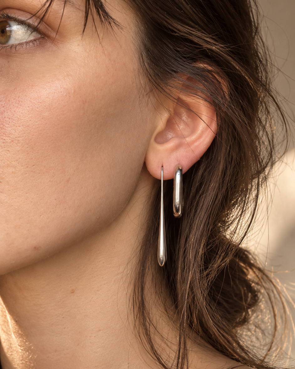 A close-up of a model's ear wearing a pair of polished silver drop hoop earrings, with her hair and skin illuminated by warm natural light.
