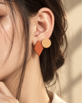 A close-up of a woman wearing a geometric stud earring with a terracotta orange diamond and a light beige circle, set against a warm beige background.