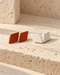 Two minimalist square stud earrings displayed on a textured beige stone surface. One earring is terracotta orange, and the other is matte off-white/cream.