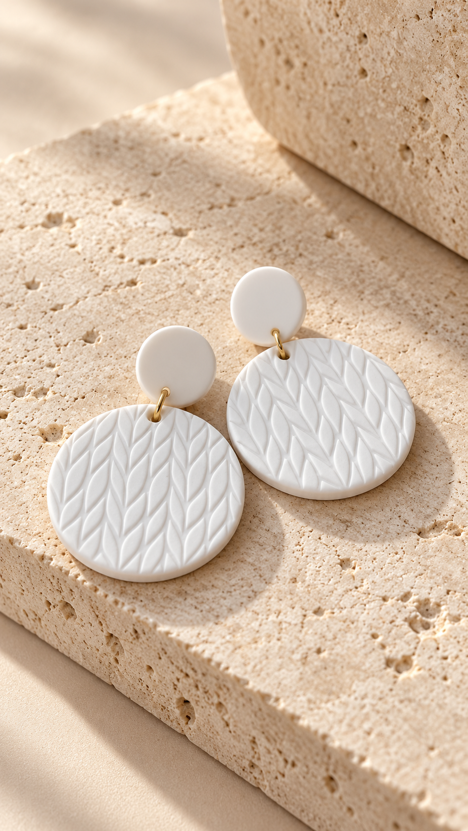 A pair of white textured disc drop earrings with small gold-tone connectors, displayed on a beige stone surface.