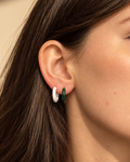 A close-up of a woman wearing a pair of small gold hoop earrings with white and green enamel details, set against a warm beige background.