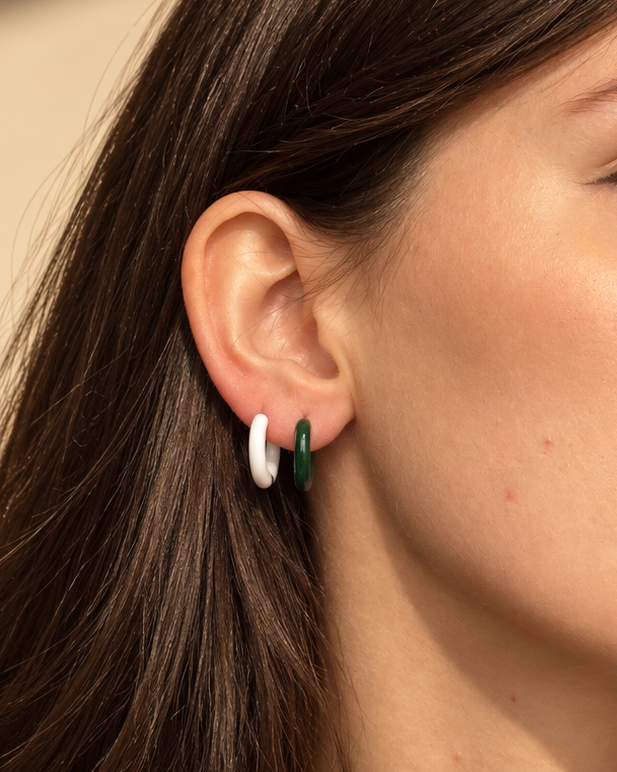 A close-up of a woman wearing a pair of small gold hoop earrings with white and green enamel details, set against a warm beige background.